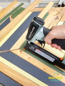 nail gun nailing down strips of painted wood onto plywood to create geometric design for twin bed frame
